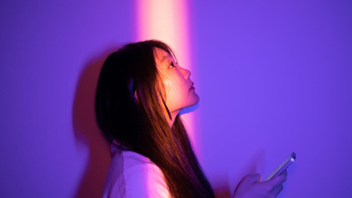 Asiatische junge Frau benutzt ihr Smartphone, während sie nachts im Scheinwerferlicht steht © Qi Yang / Getty Images